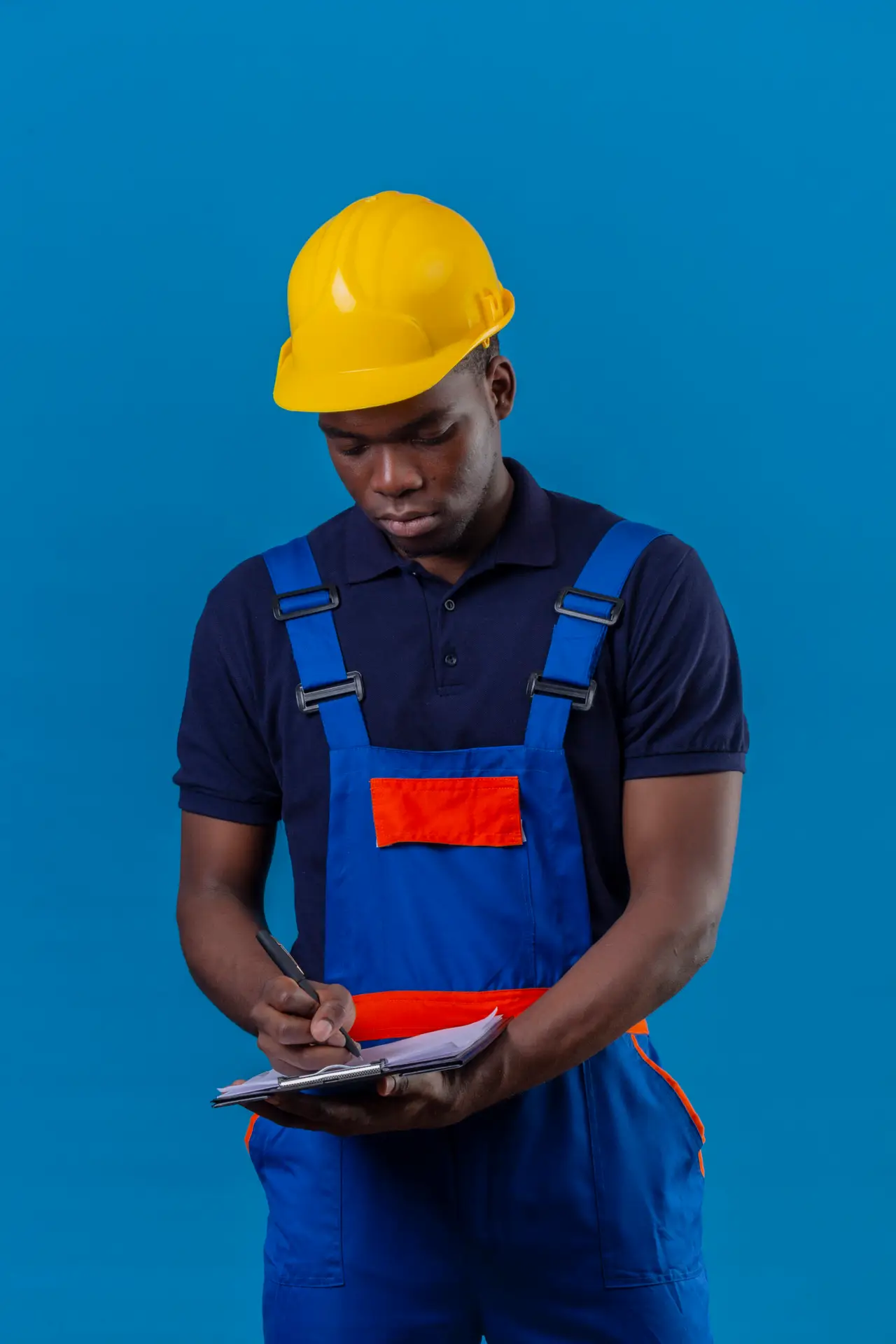 young african american builder man wearing construction uniform and safety helmet holding clipboard making notes with pen standing over blue background